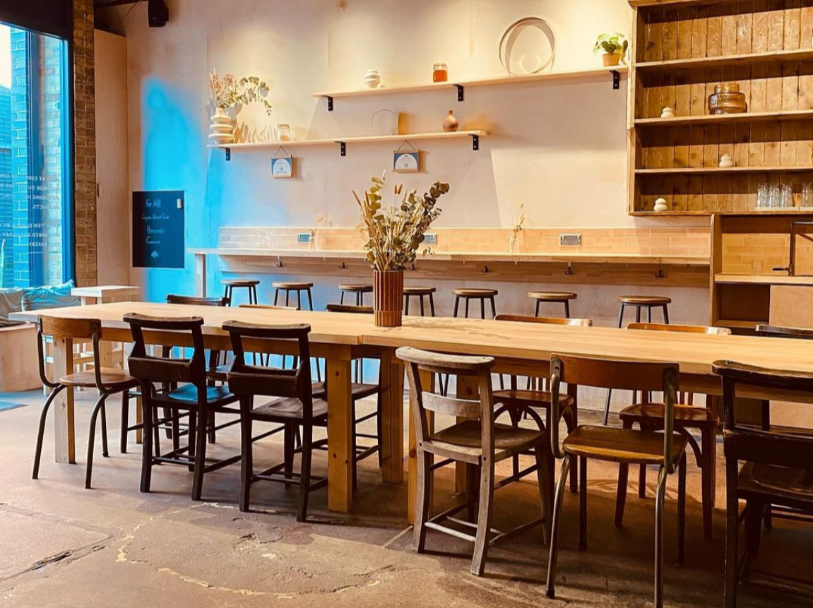 Inside of the Maltby Bakery with a large sharing table in the middle of the room and chairs around it, additional sitting against the wall with bar stools.